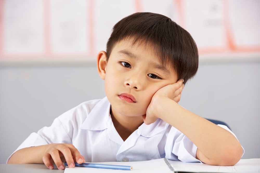 Unhappy Male Student Working At Desk In Chinese School Classroom